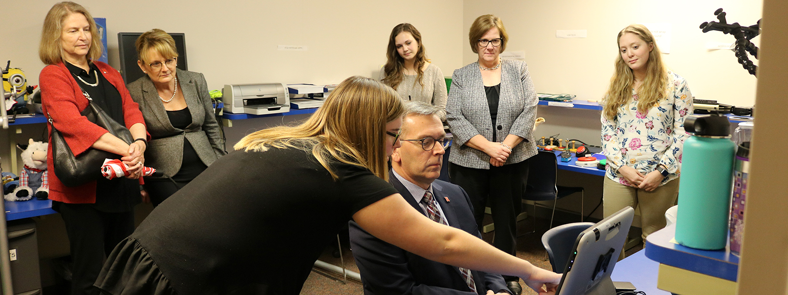 Chancellor Ronnie Green receives instructions from Abby Crimmins, a speech-language pathology graduate student, on how to use the eye gaze system in the AAC Lab as several people look on.