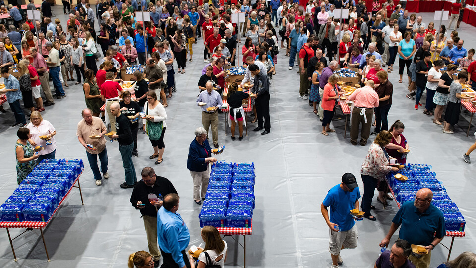 An indoor event with a large crowd. Groups of people going through a line of tables setup similar to a lunch buffet.