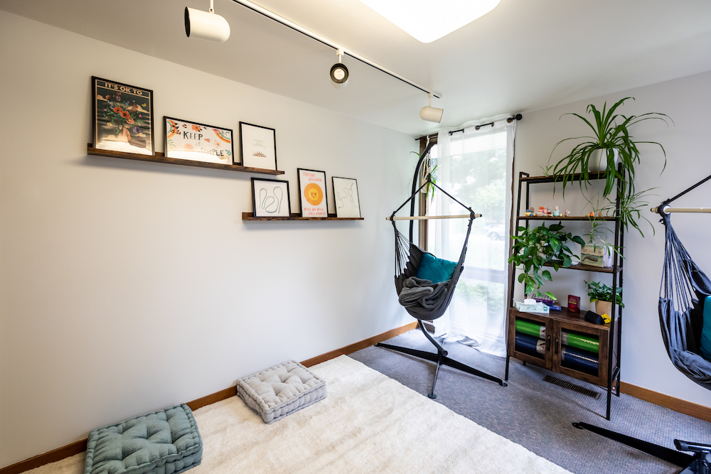 A well-lit room featuring pillows on the floor, cheerful artwork, and two hammock chairs. 