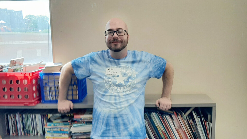 Nick Angelos wears a tie-dyed blue t-shirt and stands in front of book shelves in a classroom. 