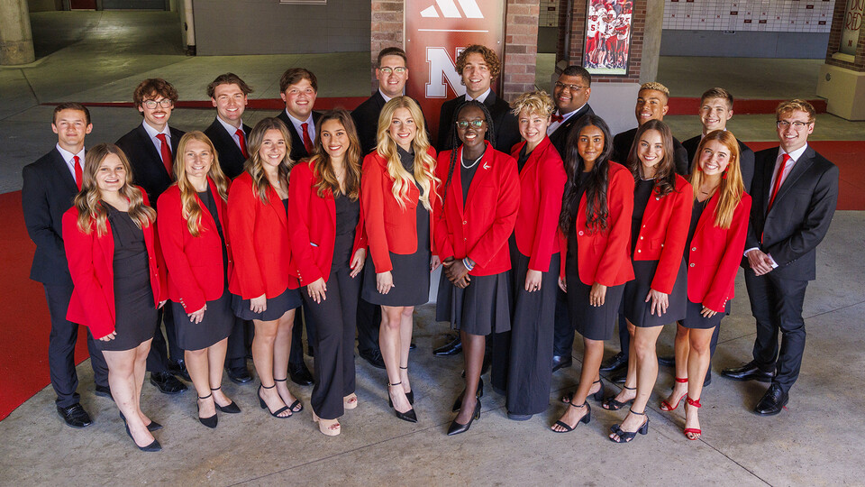 Twenty people wearing professional attire of red and black stand in front of a brick column. 