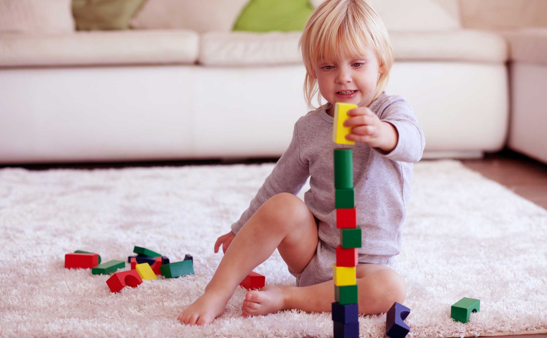 A young child with blonde hair, sitting on a light colored carpet, stacking colorful blocks. 