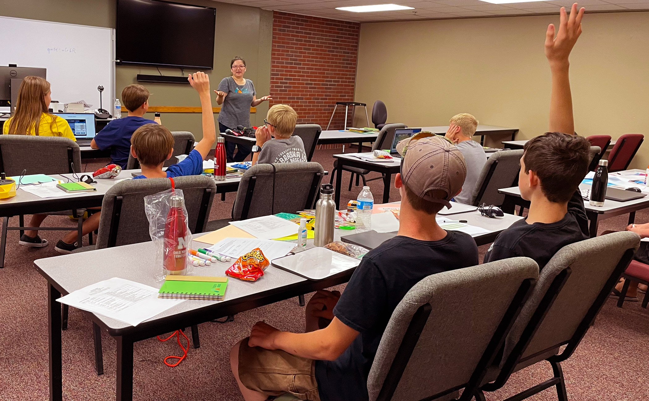 A tan and brick walled classroom setting. Instructor speaking to a group of students. Lots of school supplies and snacks sitting on tables. Photo taken from the back of the room. 