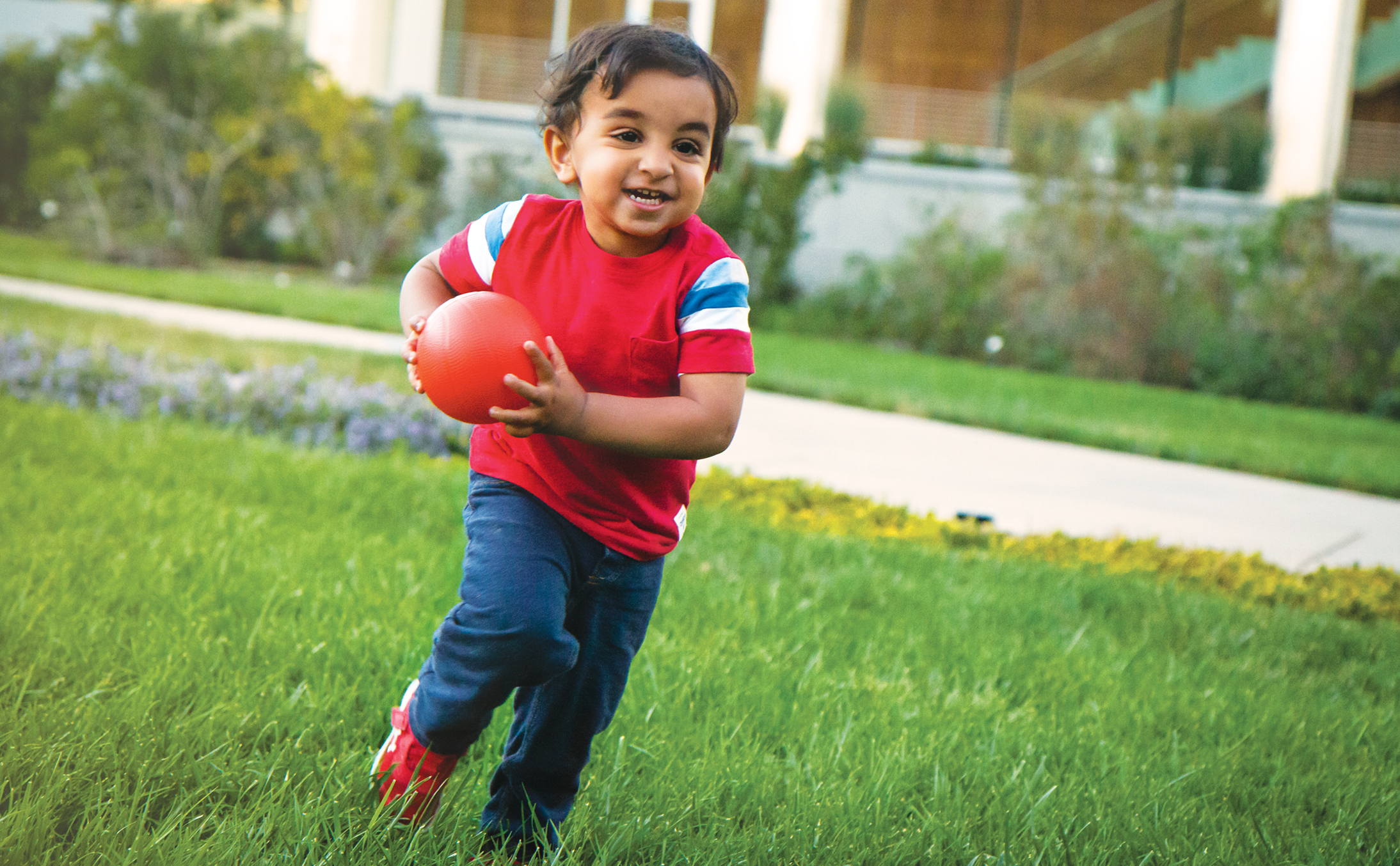 Small child playing on the grass outside. Child is wearing a red shirt with jeans, and is running with a small orange ball. 