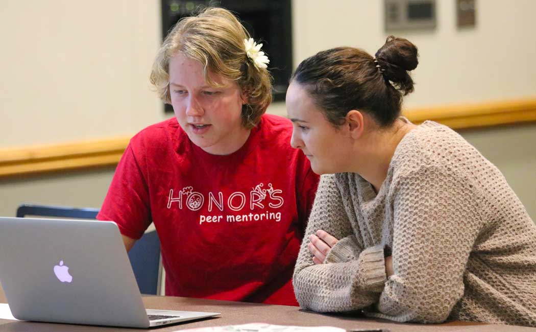 Two individuals sitting at a table looking at a computer. One is wearing a read shirt that reads "Honors peer mentoring" and the other is wearing a tan sweater.