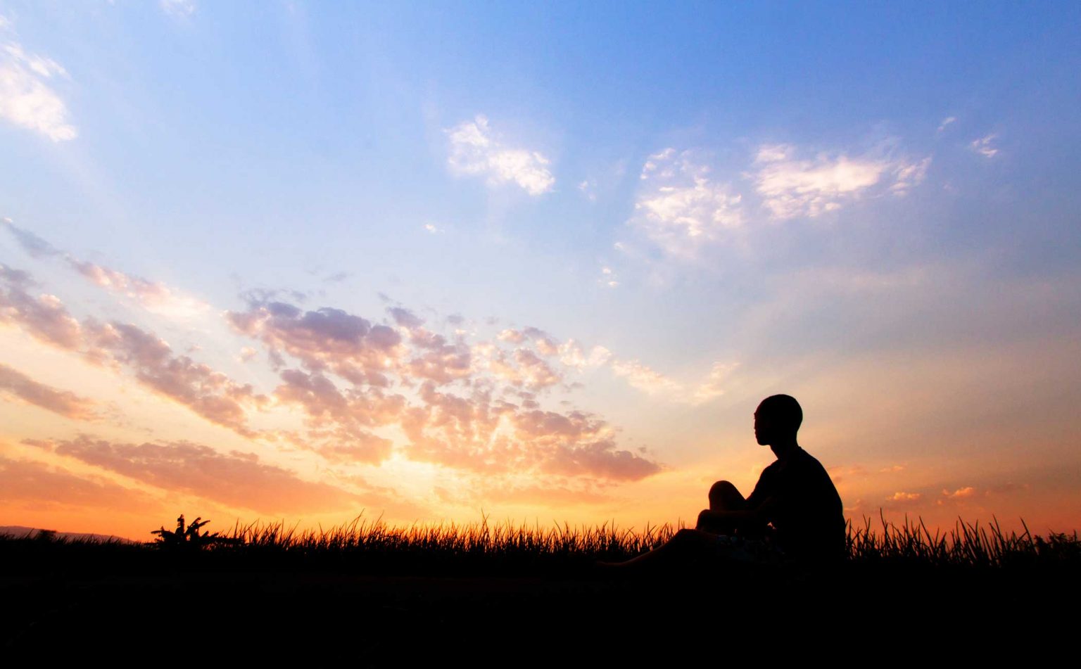 The silhouette of one individual sitting outdoors watching the sunset on top of a hill. 