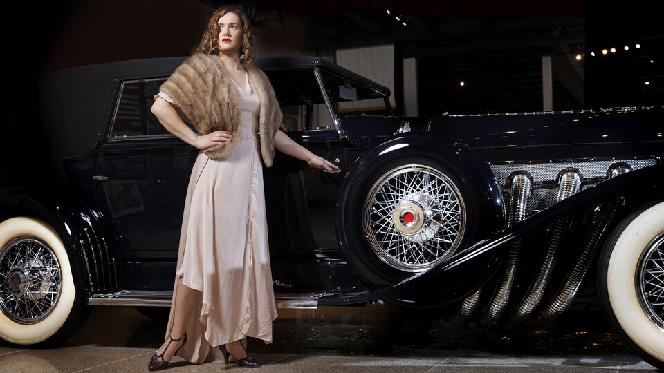 Anna Kuhlman stands in front of a 1930 Duesenberg Model J wearing pink 1930's style gown.