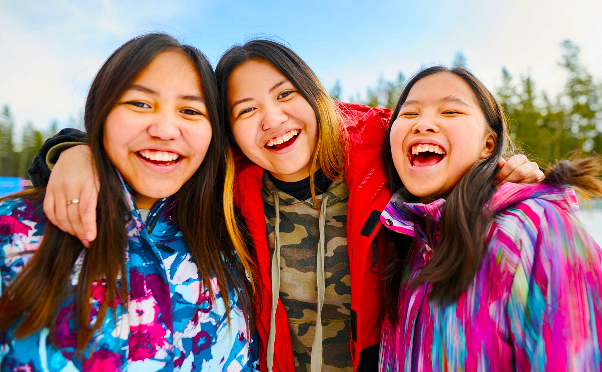 Three Indigenous youth wear bright clothing and smile for a photo while standing outdoors. 