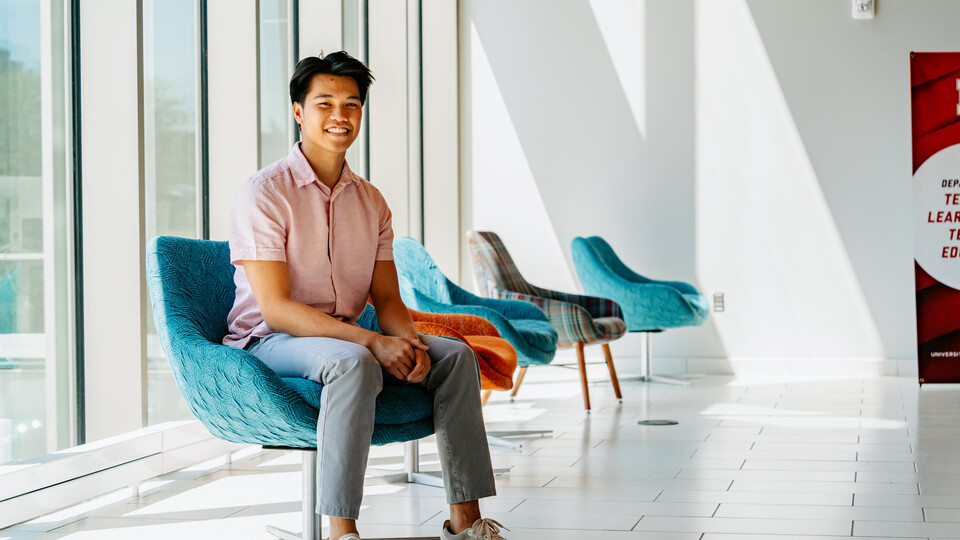 Andre Nguyen sits in a blue chair along a wall of windows in Carolyn Pope Edwards Hall. 