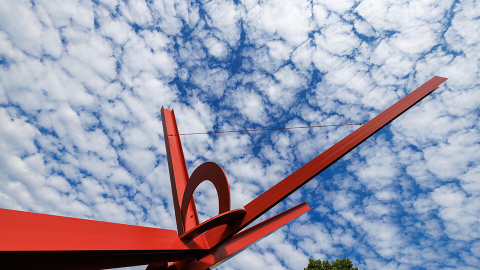 Red sculpture photographed upwards with blue  sky and clouds behind. 