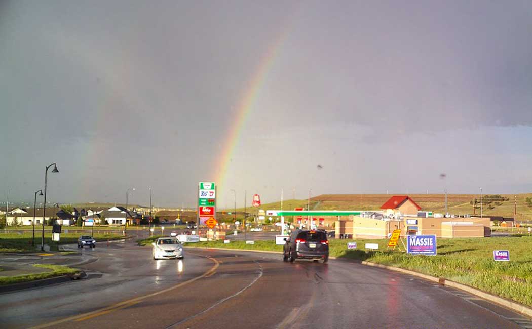 A rainbow shines over a highway with a gas station and other community buildings on each side. 