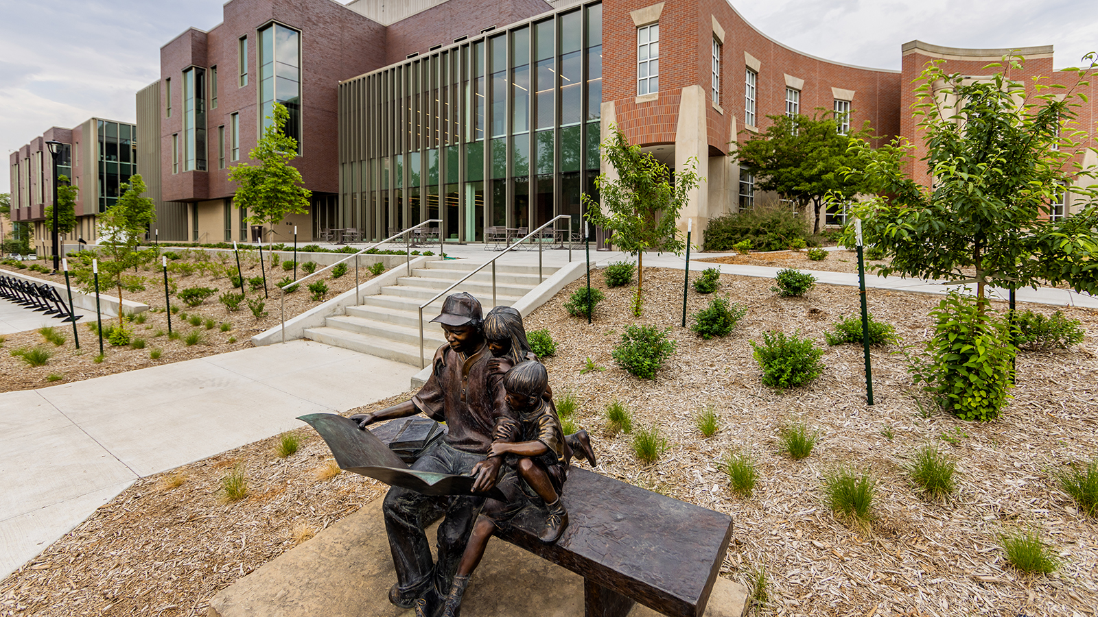 sculpture of man reading a book to two children on a bench with Carolyn Pope Edwards Hall and Teachers College Hall in background