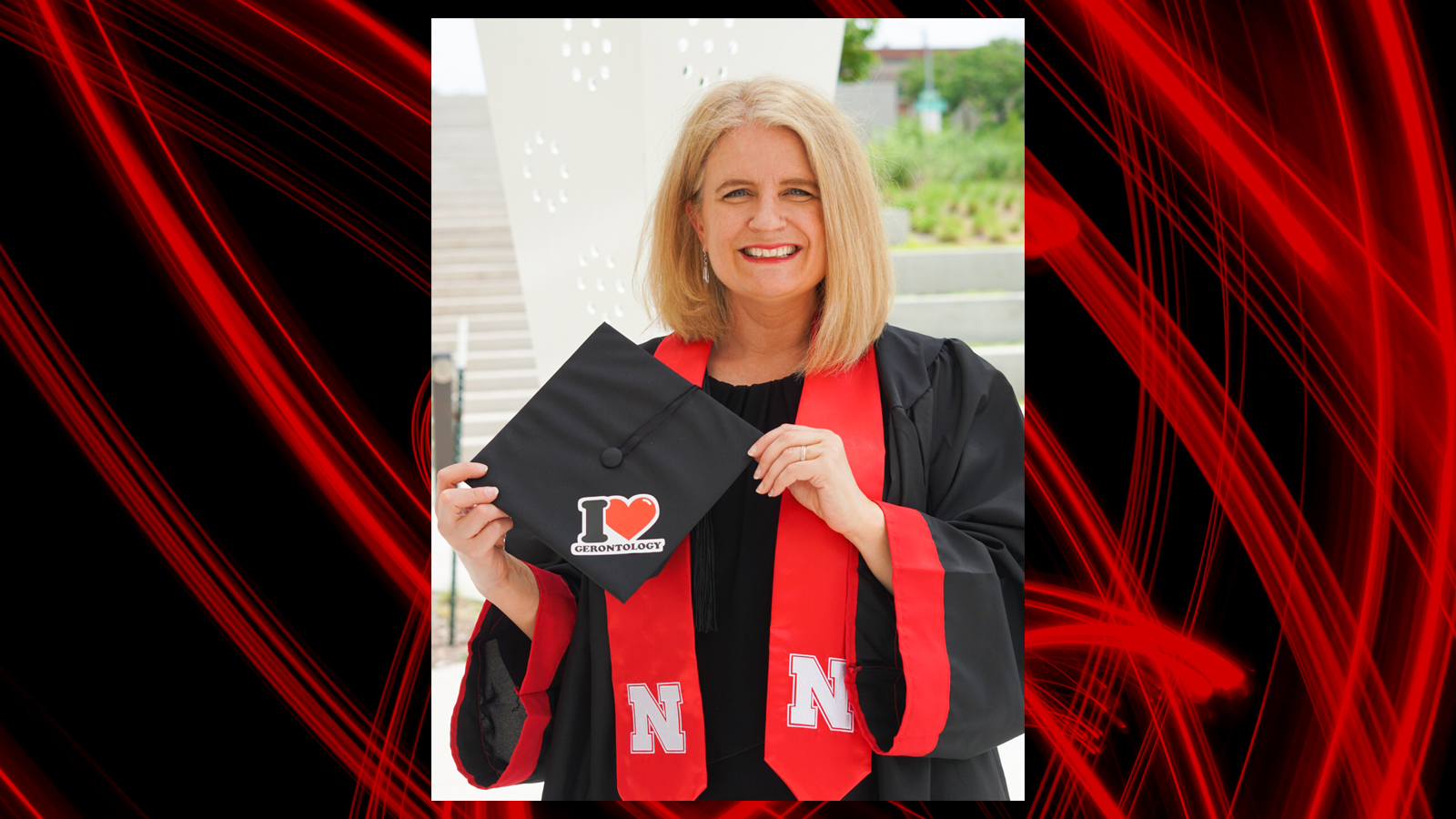 Kristin Johnson poses in her graduation regalia holding her mortar board that has a "I heart gerontology" sticker; black background with red brush strokes