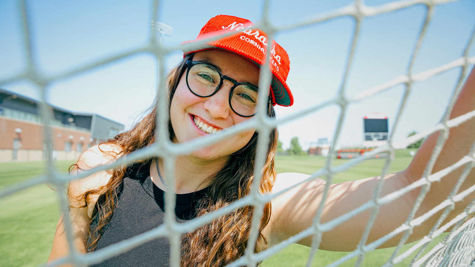 Cece Villa smiles through a soccer net while wearing a red Nebraska cap and black tank top; photo by Kristen Labadie | University Communication and Marketing