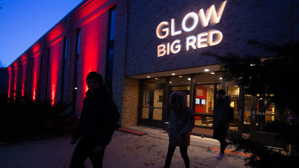 Photo at nighttime of a brick building lit up red. A light projects "Glow Big Red" above the entrance doors. 