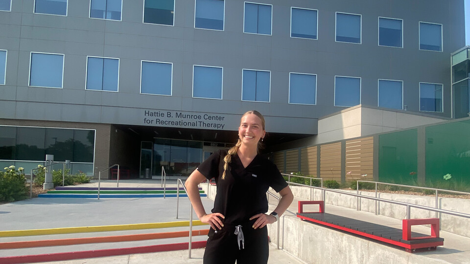 Elsa Wilcox wears black medical scrubs and stands in front of a large building - the Hattie B. Munroe Center for Recreational Therapy. 
