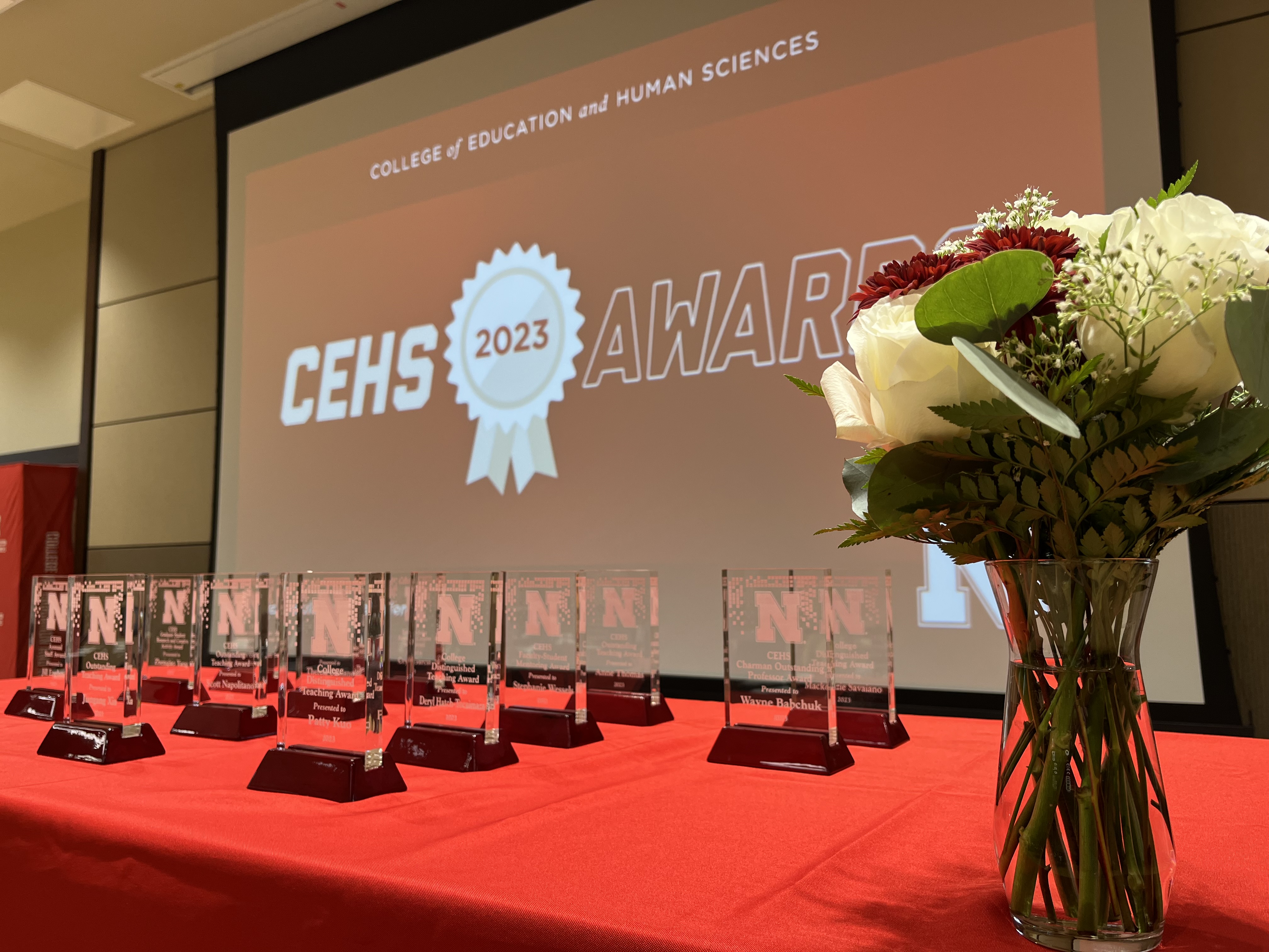 A table with a red tablecloth, awards, a bouquet of red and white flowers in front of a screen that reads CEHS 2023 Awards