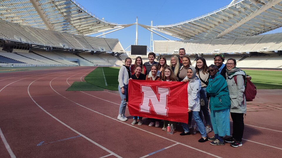 A large group of students stand in an outdoor stadium holding a red flag with an N on it. 