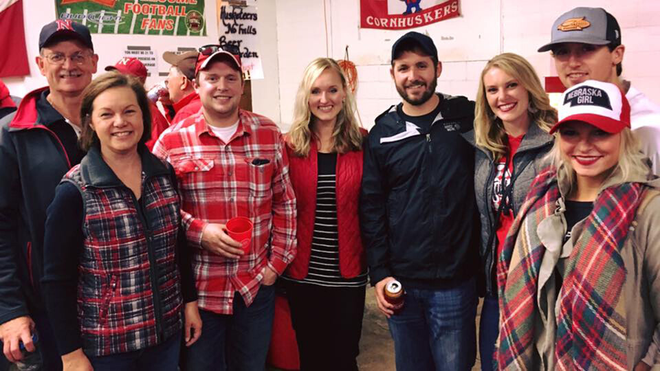Eight people dressed in red and black smile for a photo at a Husker gathering. 