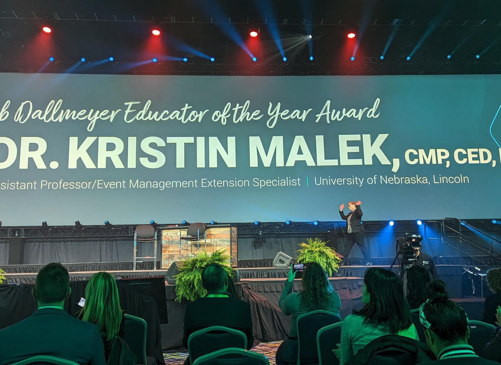 Kristin Malek walks across a large stage with a screen displaying the educator of the year award information. 