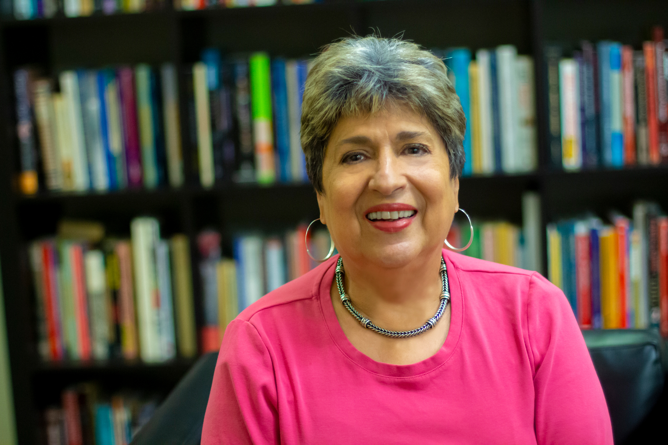 Ofelia Garcia, smiles for a photo while wearing bright pink clothing and sitting in front of book shelves. 