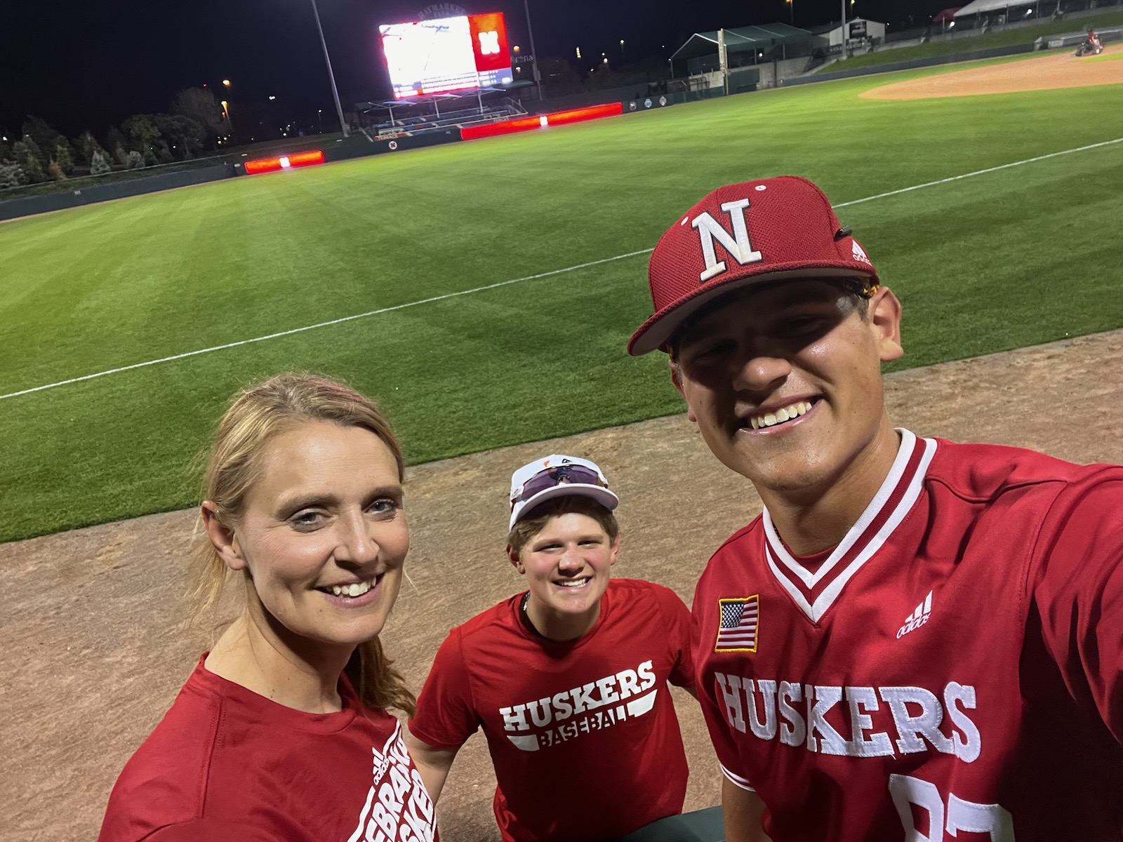 Three people, wearing red, smile for a photo with the green grass of a baseball field behind them. 