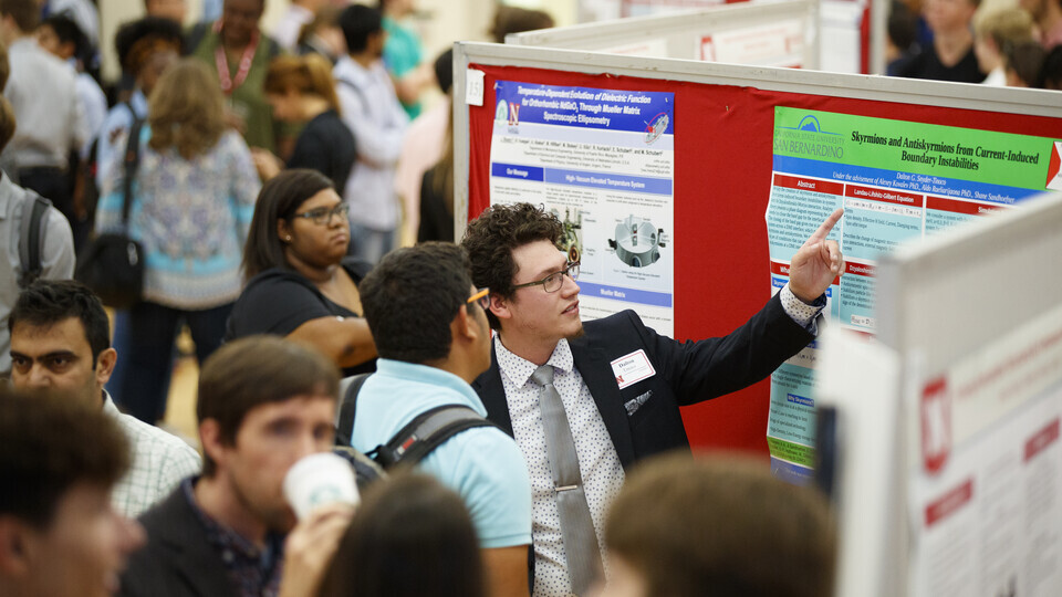 One student dressed in a student, points to his research poster during a well-attended research fair. 