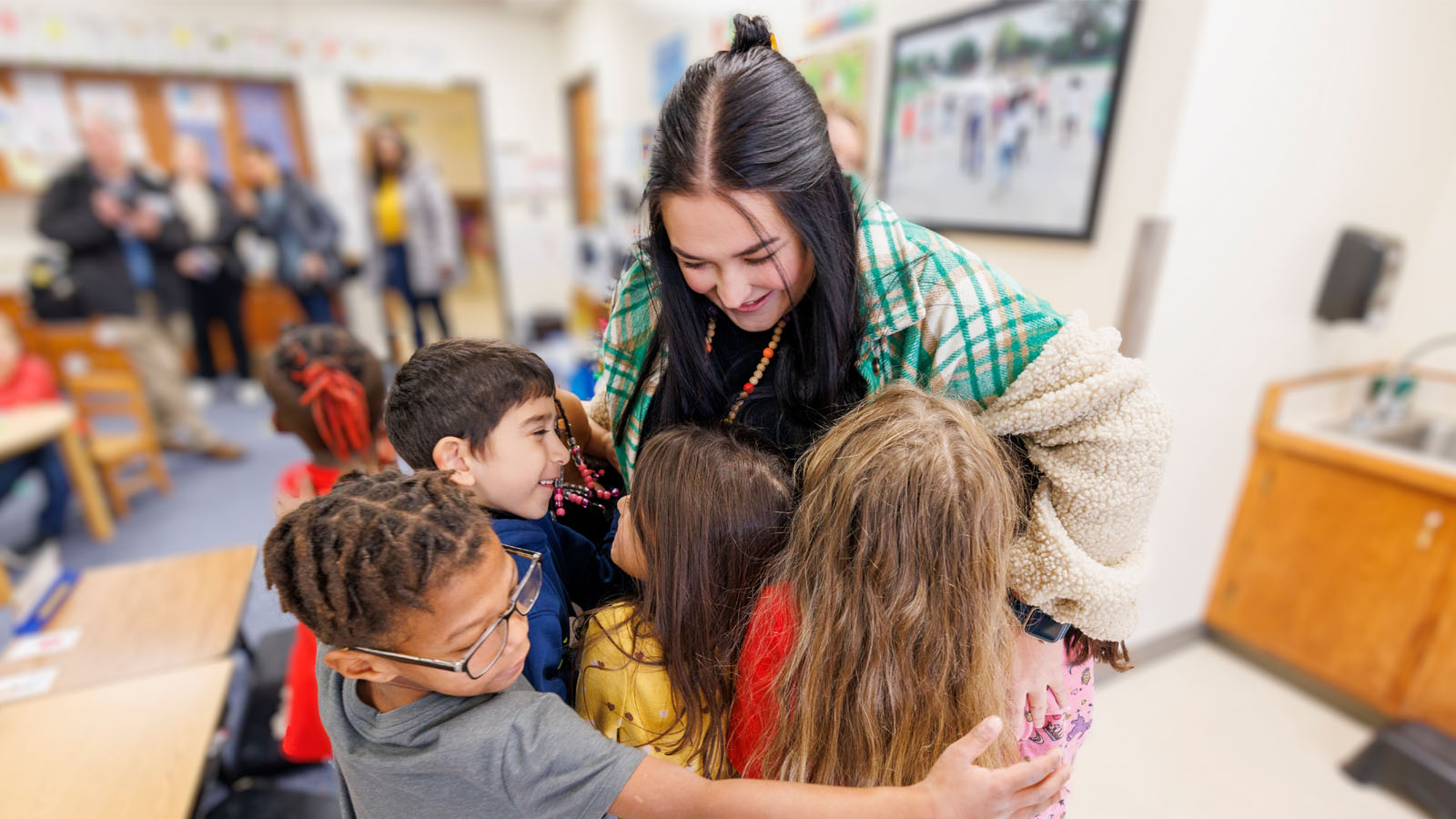 Teacher hugging a group of young children in a brightly lit classroom setting.