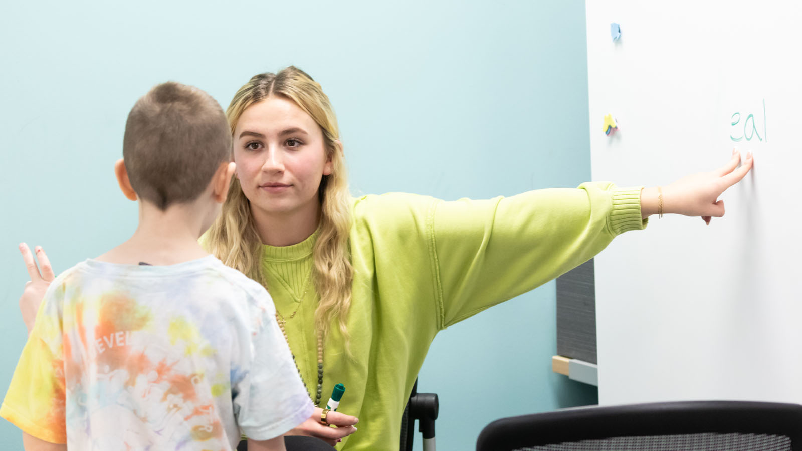 A teacher points to a whiteboard while a child listens attentively in a classroom setting.