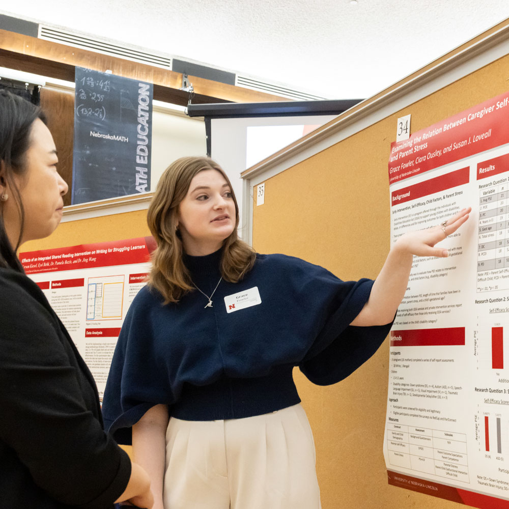 Person presenting research poster, gesturing to data while another listens attentively.