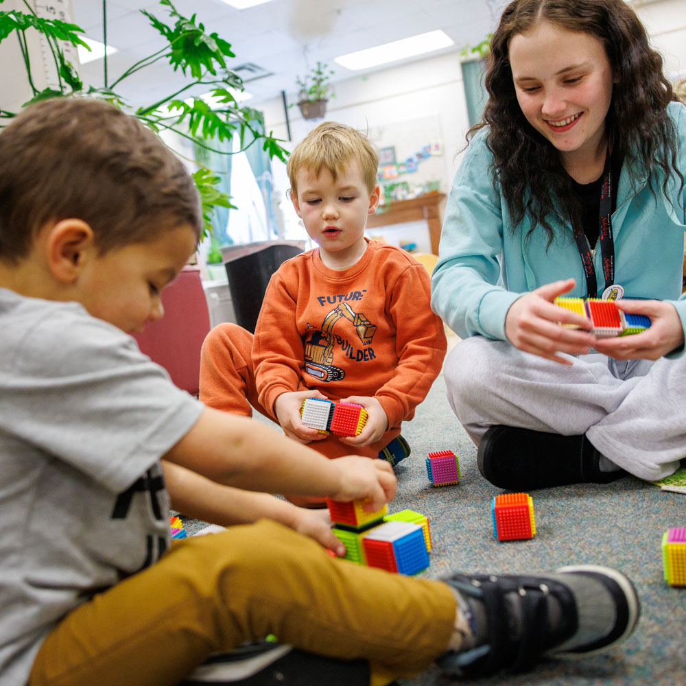 Children and adult sitting on the floor playing with colorful cube toys in a bright room.