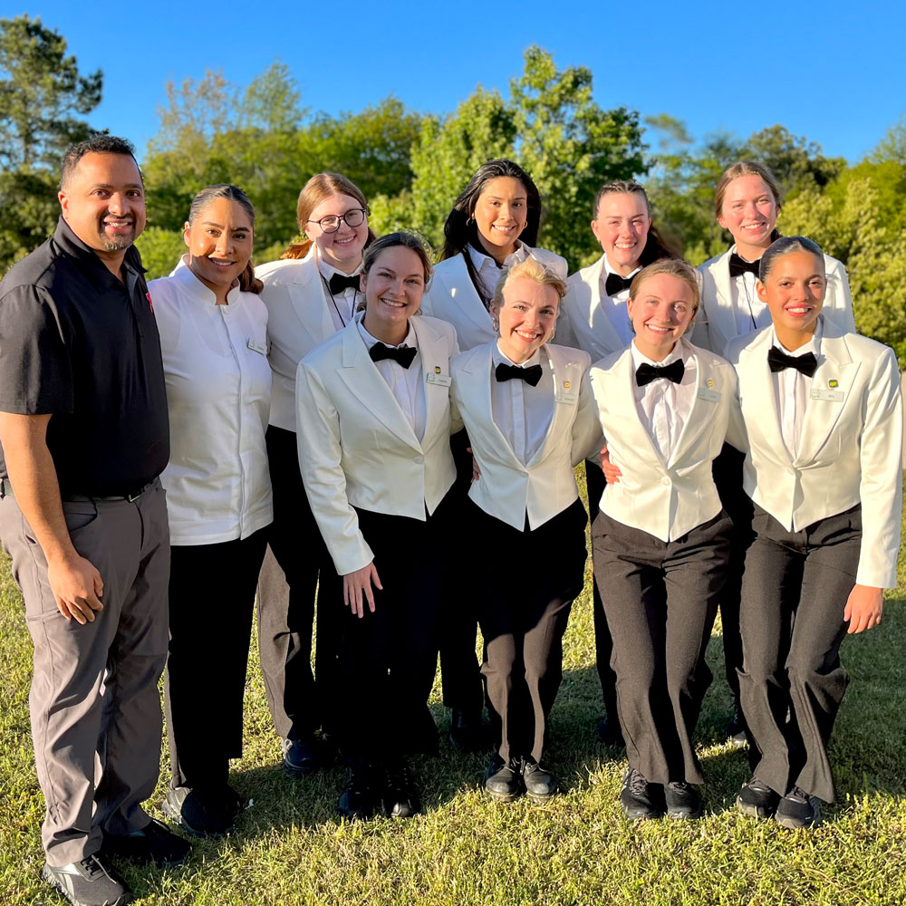 Group of people outdoors, wearing formal uniforms with black bow ties, standing on grass with trees behind.