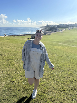 Natalia Vazquez poses for a photo on a hillside with the ocean and a city far in the background