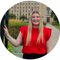 Smiling woman (Abbie Leu.) in a red blouse standing outdoors near a building and greenery.