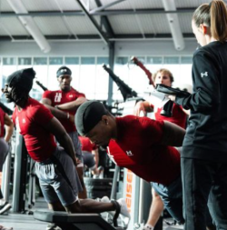 Group exercise session in a gym, participants in red shirts stretching, trainer observing.