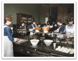 Women working in a kitchen, preparing food on long tables, wearing aprons and hats.