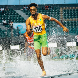 Track athlete running through water in a steeplechase race, wearing green and yellow, mid-action.