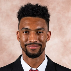 Man in a suit with a red tie, smiling in front of a beige backdrop.