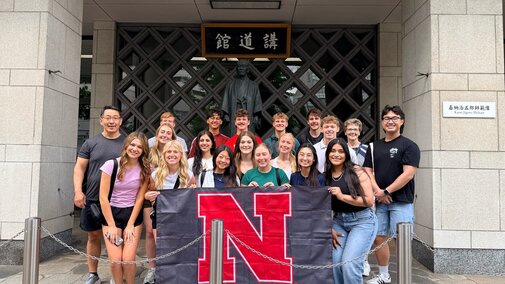 Group of people smiling, holding a banner with a large red "N" in front of a building.