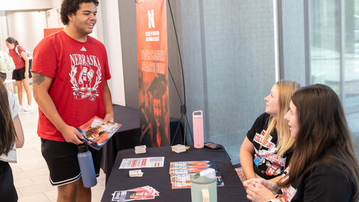 Student at a table talking to two seated representatives, holding brochures in a bright indoor space.