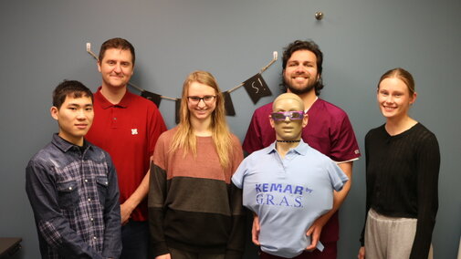 William Salloom and his students pictured with a mannequin body. The backdrop is a blue and gray mixed wall with a strigned banner hanging on it.