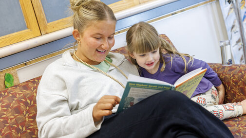 Woman and young girl reading together on a patterned sofa, both smiling and engaged.