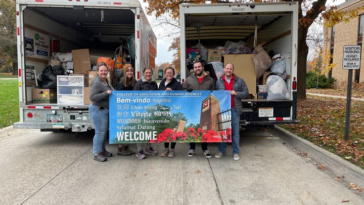 Six people hold a sign in front of two moving trucks full of donated items. Outdoor photo. 