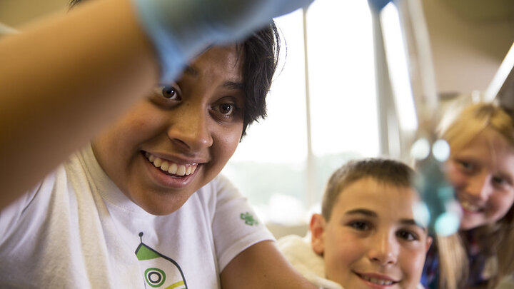 An adult wearing a white t-shirt and blue gloves conducts a scientific experiment with two youth. 