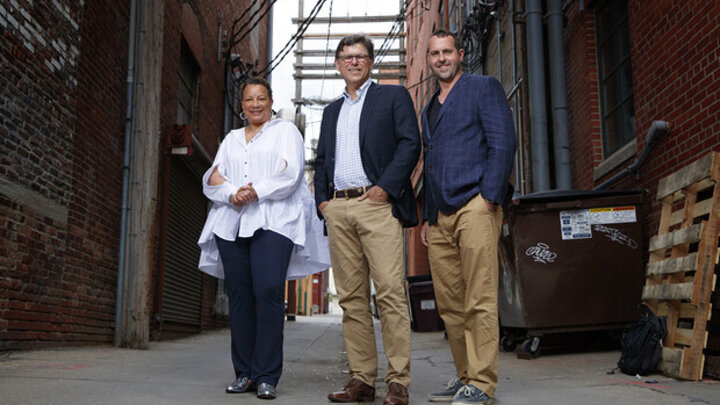 Three individuals standing in an ally in between two red brick buildings, with several power lines overhead. 