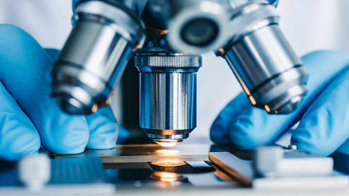 Two hands wearing blue gloves place an object underneath of a microscope. 