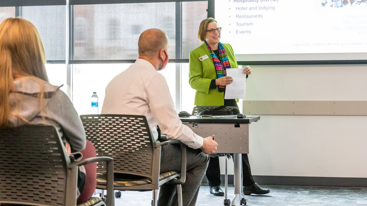 Mary Ann Johnson wearing a bright green suit jacket stands at the front of the room and speaks to people sitting in chairs. 