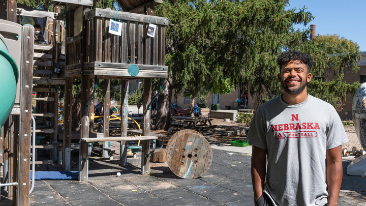 Outdoor scene. Adult individual wearing a gray t-shirt stands near a playground and poses for a picture. 