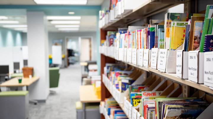 Books line the shelves at the Schmoker Reading Center. In the backgorund you see an office setting with glass walls and computer desks. 