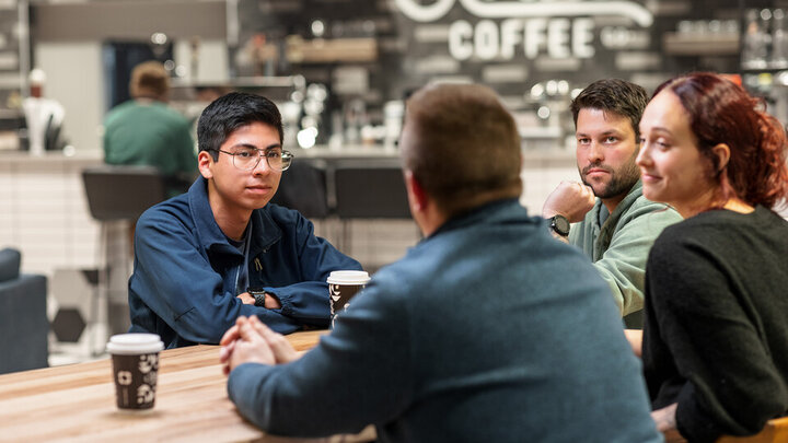 Four individuals sit around a wooden table, drinking coffee, engaged in conversation. 